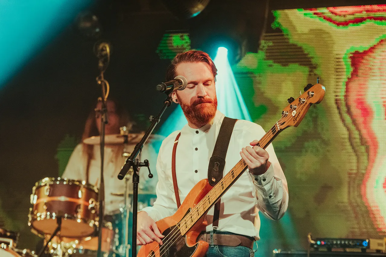 Bearded musician playing electric bass guitar on stage with microphone in front, colorful lights in background.