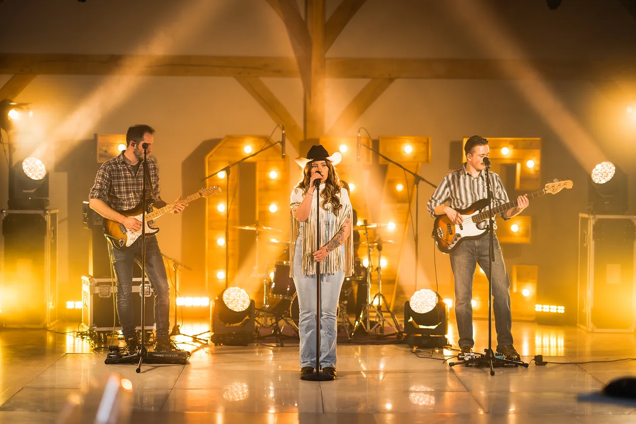 Three musicians performing on stage with warm yellow lighting; a female singer in a cowboy hat stands center, flanked by two male guitarists.