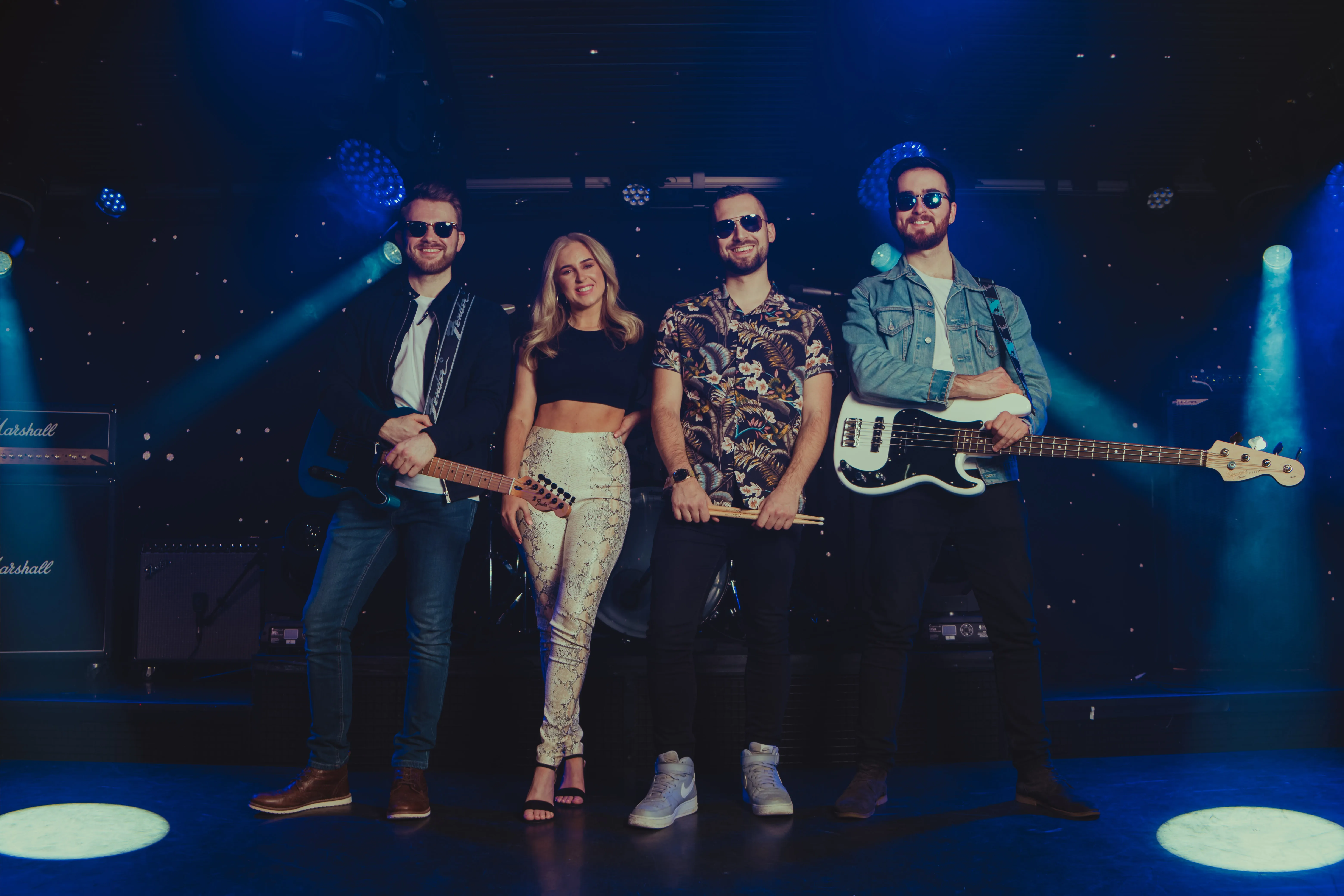 Four musicians wearing sunglasses stand on a stage with instruments and blue spotlights in the background.