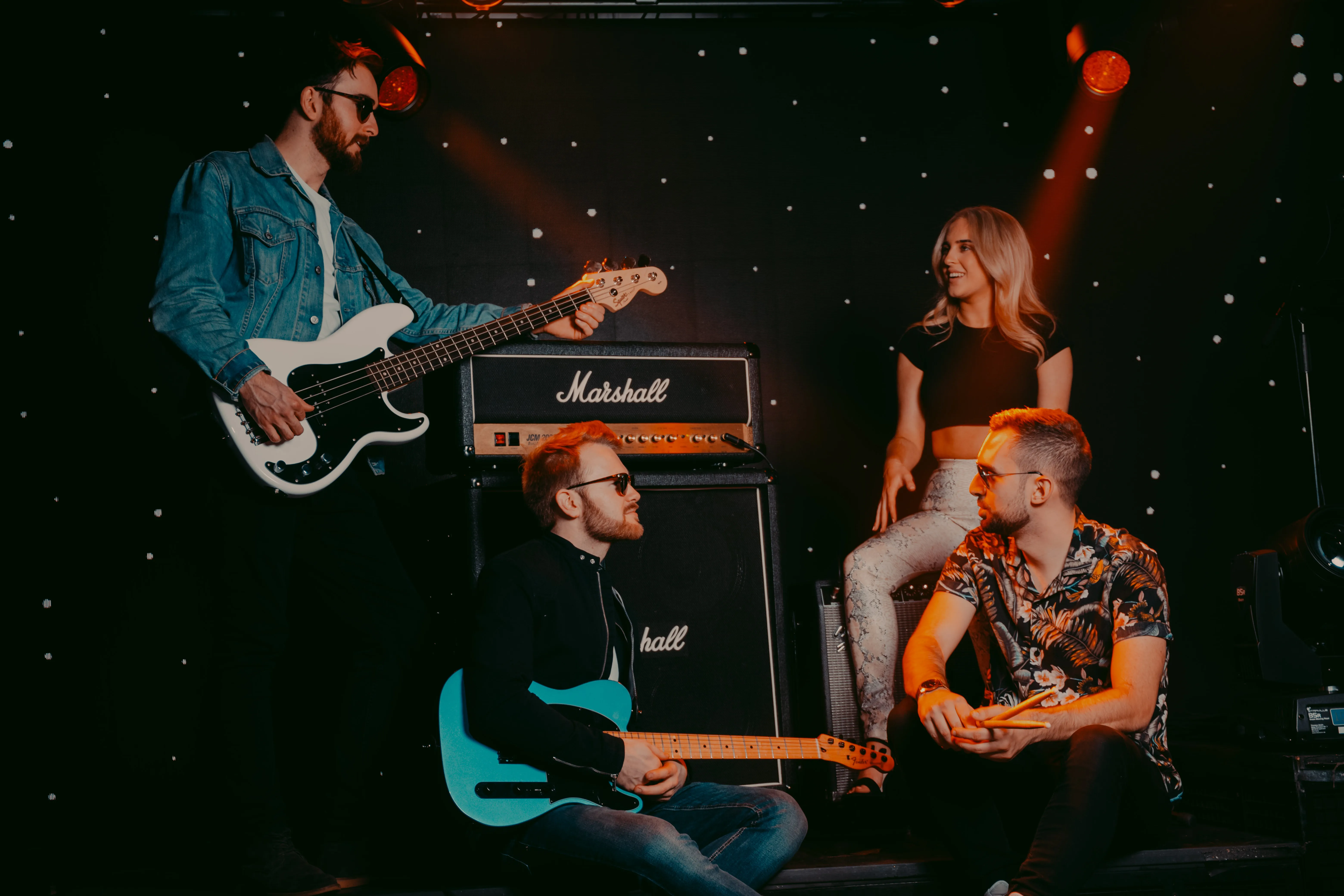 Four musicians with guitars and drumsticks in a dimly lit room with Marshall amplifiers and star-like lights in the background.