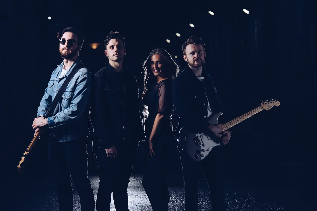 Four band members standing on a dark street at night, two men holding electric guitars, one woman in the center with long hair.