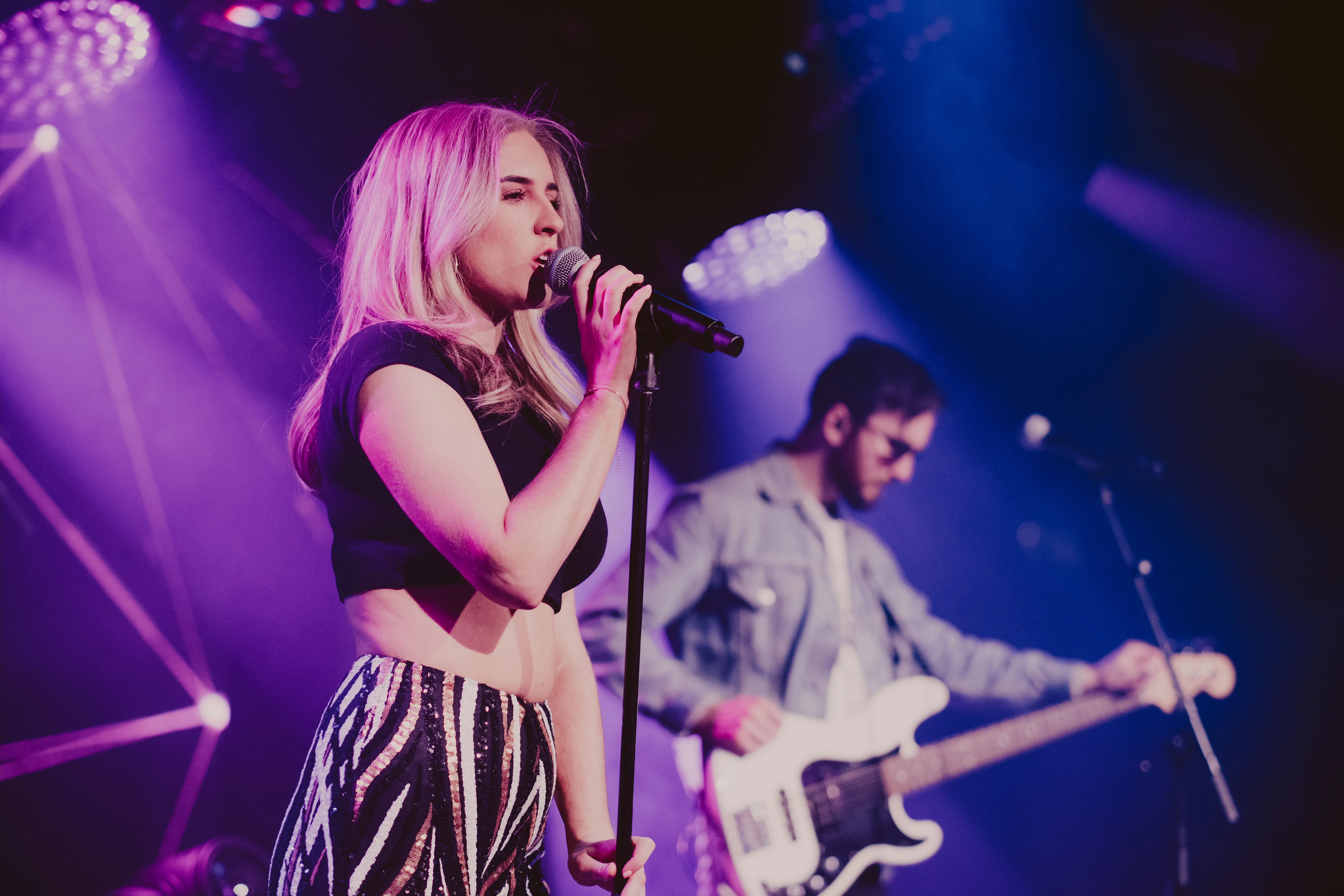 Female singer with long blonde hair performing on stage with a microphone, and a male bassist playing beside her under purple and blue stage lights.