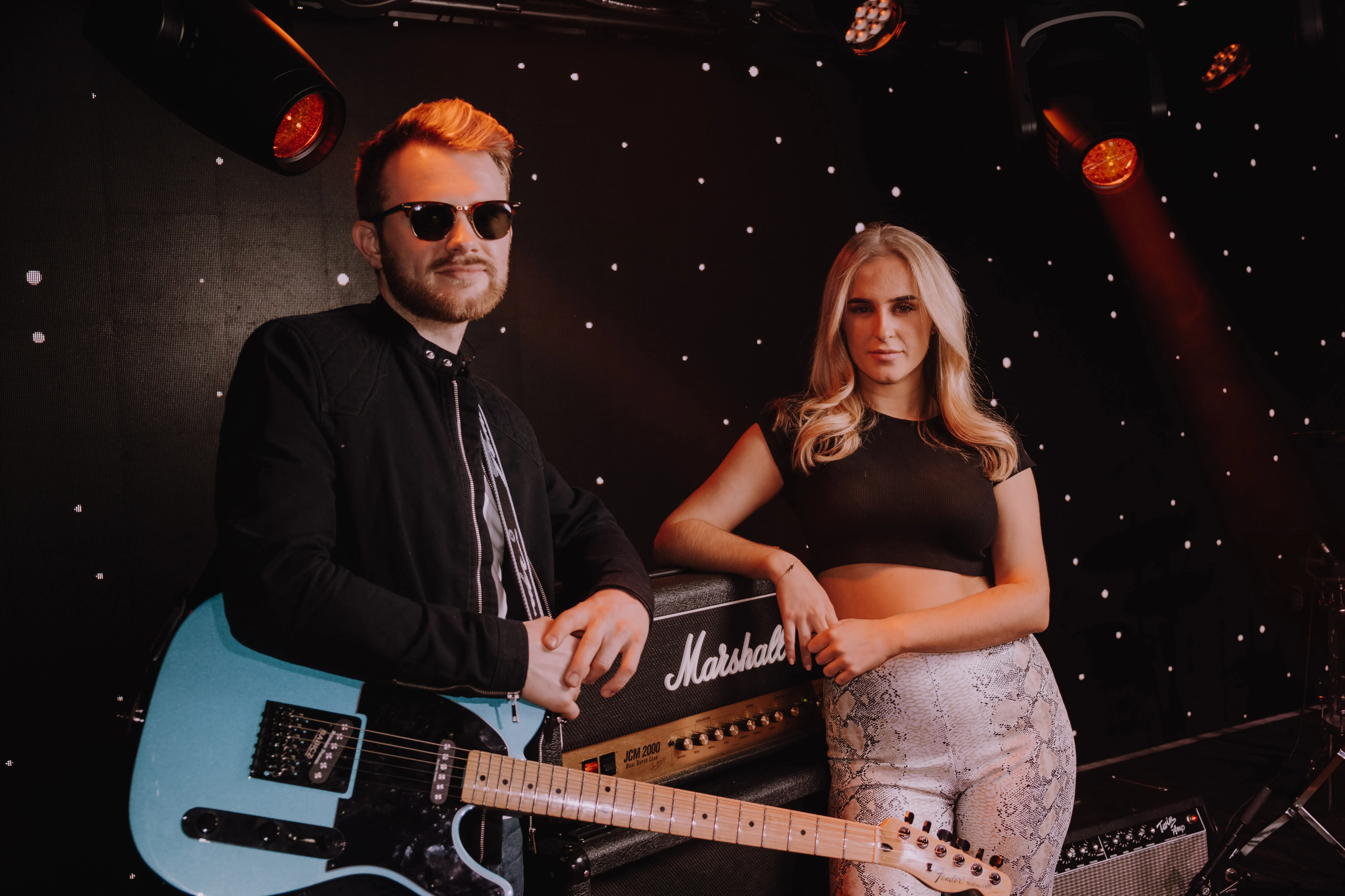Man wearing sunglasses and black jacket holding a light blue electric guitar stands next to a woman in a black crop top and patterned pants in front of a Marshall amplifier on a dark stage with star-like lights.