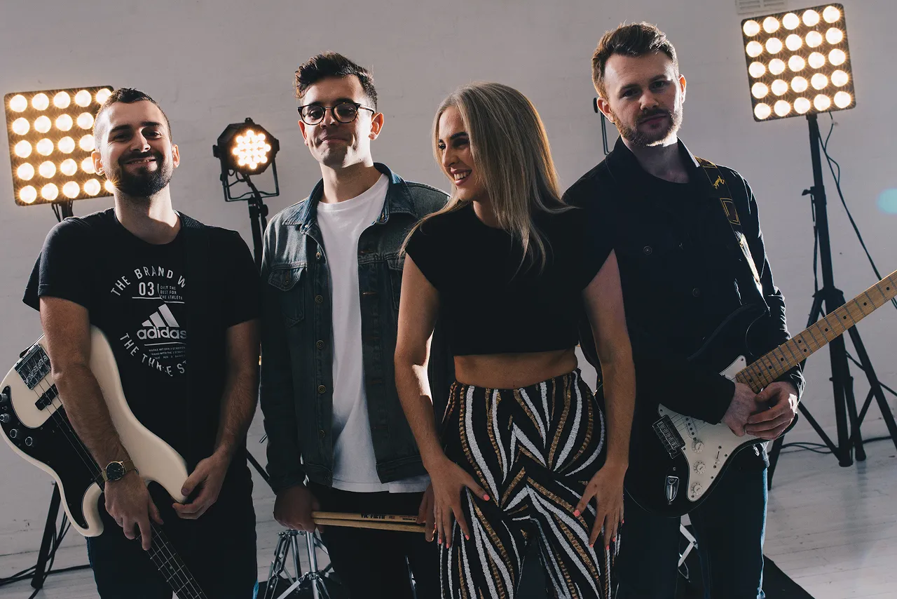 Four musicians standing in a studio with bright lights behind them, two holding guitars and one holding drumsticks.