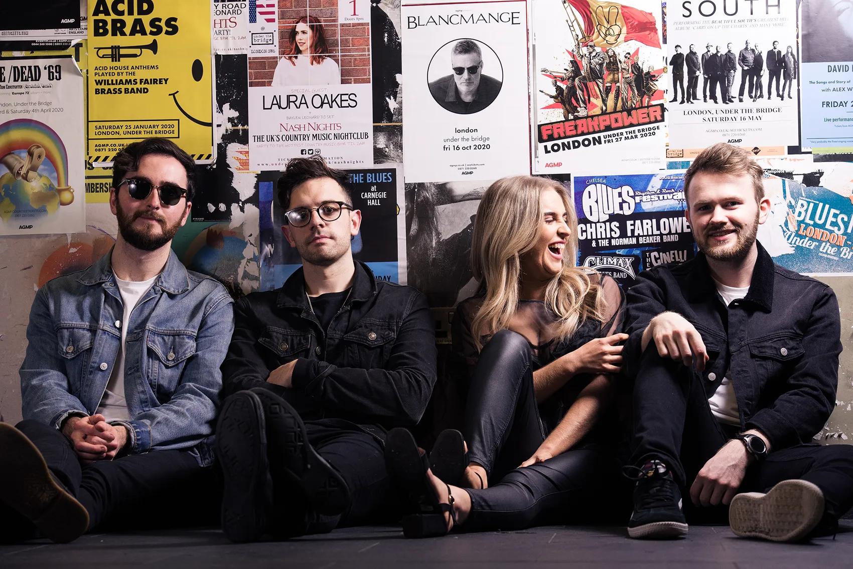 Four young adults sitting on the floor against a wall covered with colorful music event posters, two men on the left wearing glasses and two people on the right, a woman laughing and another man smiling.