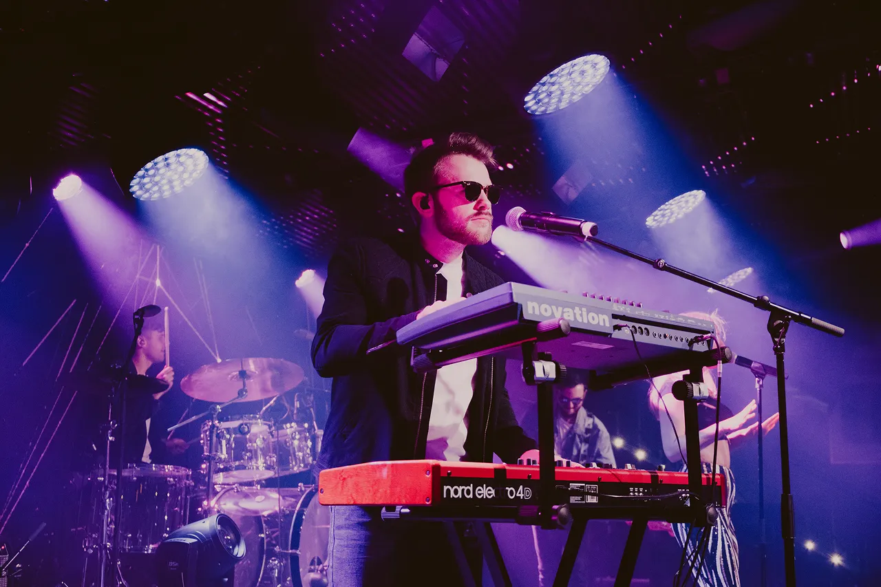 Male musician wearing sunglasses plays keyboard on stage with purple lights and drummer in the background.