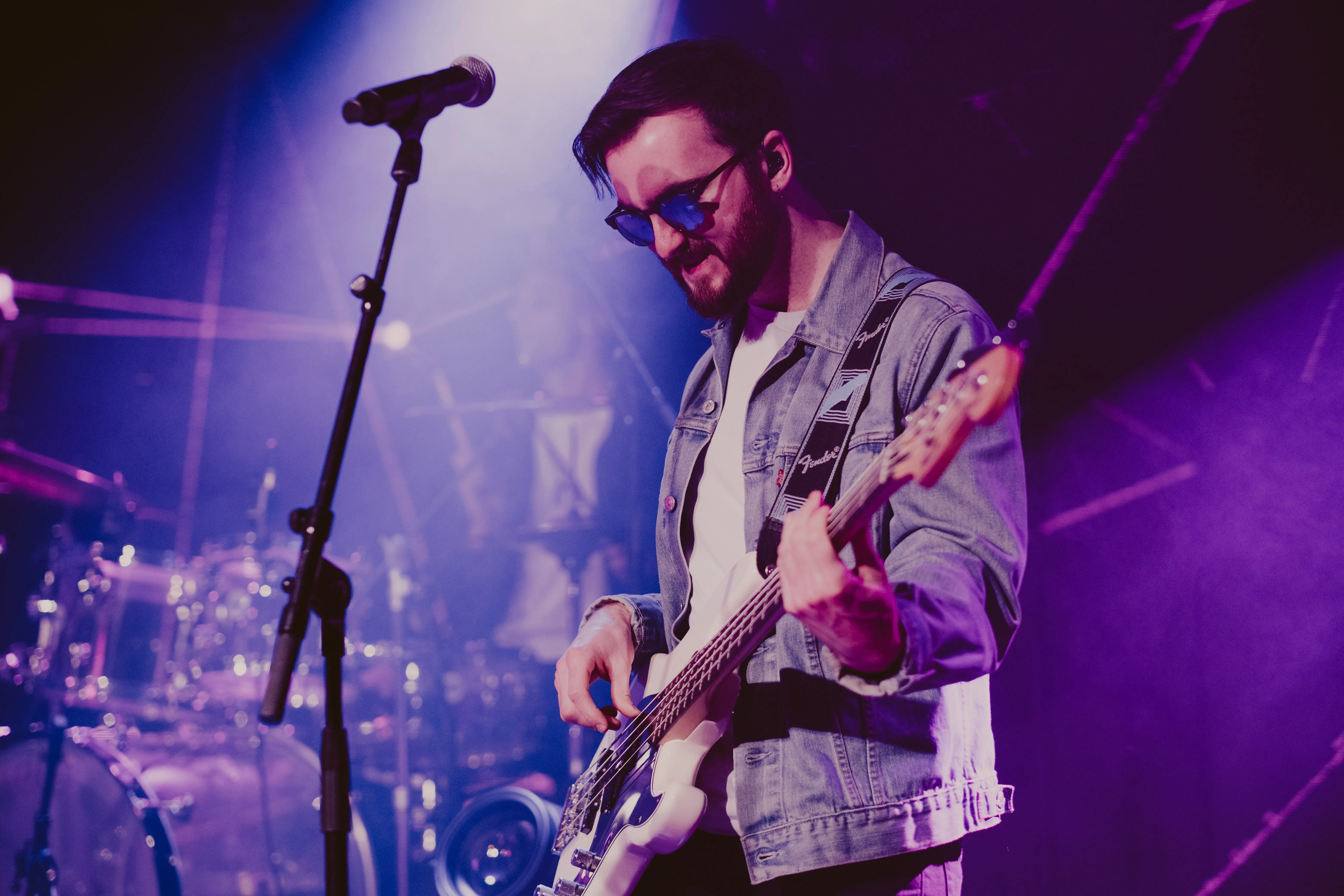Bearded man wearing sunglasses and a denim jacket playing a white electric bass guitar on stage with purple lighting.