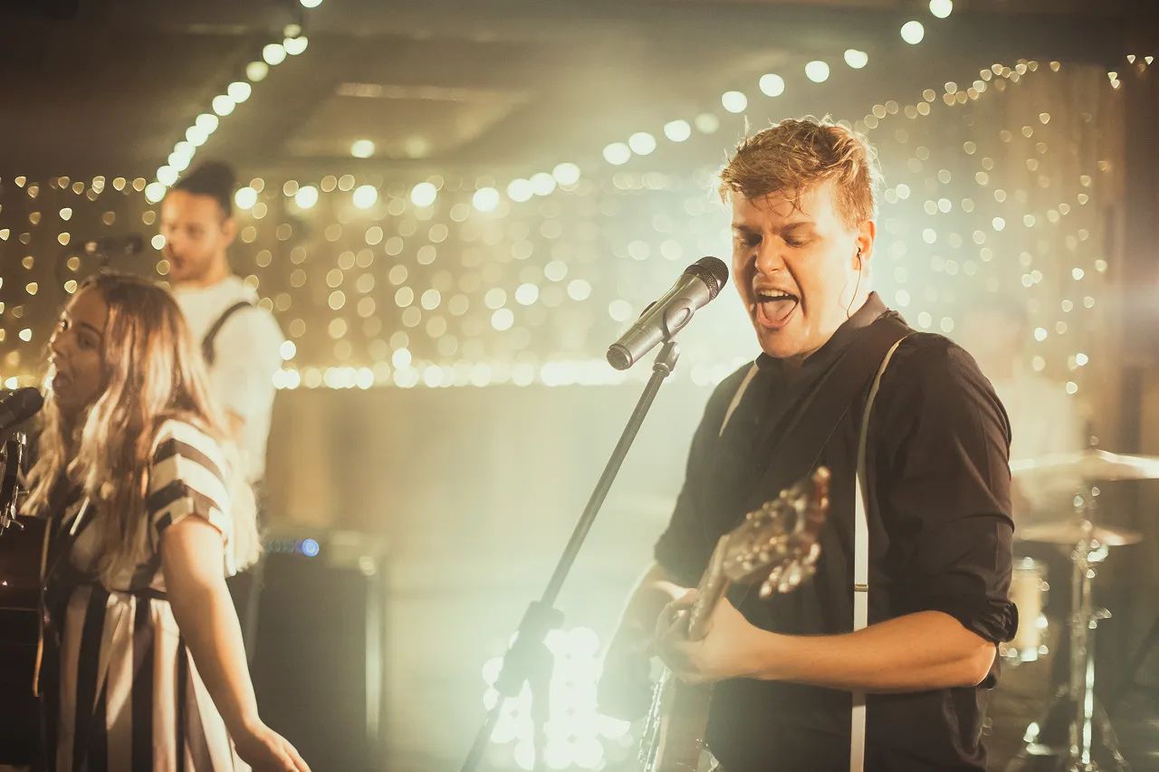 A male guitarist passionately singing into a microphone with a female vocalist and bassist performing in a softly lit room with string lights.