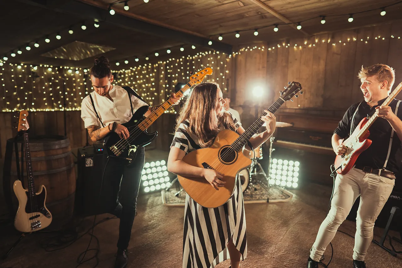 Group of three musicians playing electric bass, acoustic guitar, and electric guitar in a warmly lit room with string lights.