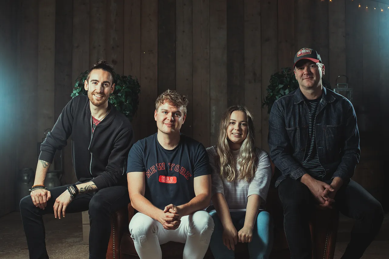 Four people sitting indoors against a wooden wall, smiling at the camera, with two men on the ends and a man and woman in the center.