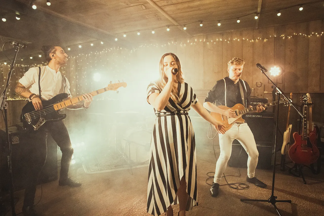 Band performing indoors with a female singer in a black and white striped dress, a bassist, and a guitarist under string lights.